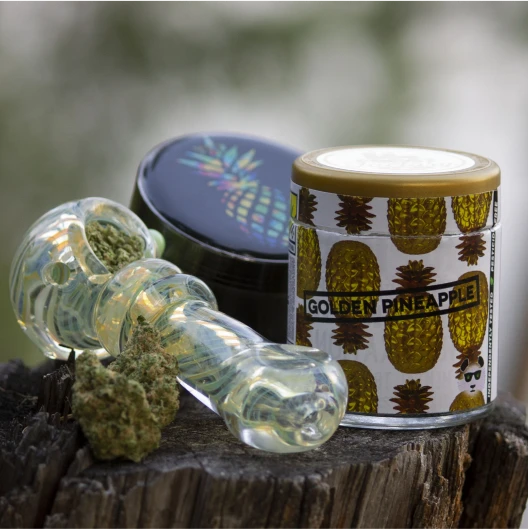 A cannabis pipe filled with ground bud sits on a bench in front of two grinders